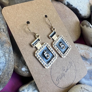 May include: Gold-coloured earrings shaped like perfume bottles, featuring black and silver details and the number 5. The earrings are adorned with small, clear crystals and hang from gold hoops. They are presented on a brown card.