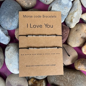 May include: Two black string bracelets with silver beads spelling out "I Love You" in Morse code on a brown card with the text "Morse code Bracelets" and "Morse code bracelet with a hidden message".