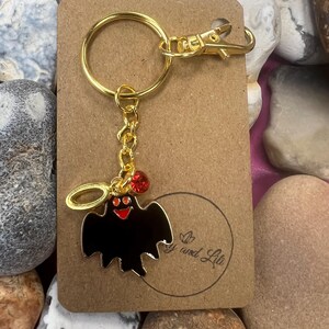 以下が含まれることがあります： 黒い幽霊のチャームと赤いラインストーンが付いたゴールドのキーチェーン。チャームは赤い目と赤い笑顔の白い顔をしています。キーチェーンはゴールドの留め金とゴールドのリングが付いています。タグには「喜びと人生」と書かれています。