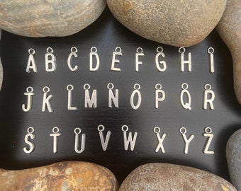 May include: Silver alphabet charms displayed on a black background, framed by stones. The set includes letters A to Z, with some letters having special characters. Suitable for jewellery making or custom crafts.