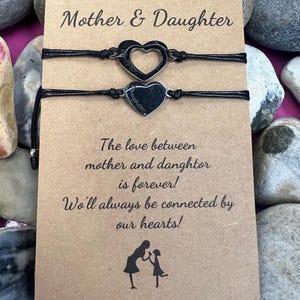 Puede incluir: Dos pulseras a juego de cordón negro con dijes de corazón plateado, presentadas en una tarjeta marrón. La tarjeta dice "Mother & Daughter" e incluye un mensaje sobre el vínculo entre una madre y su hija. El fondo presenta piedras grises lisas.