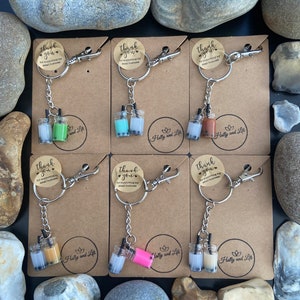 May include: Nine keychains with miniature boba tea cups. Each keychain has a small tag that says "Thank you for supporting my small business." The keychains are arranged in three rows of three. The boba tea cups are filled with different colors of boba tea, including brown, pink, green, and white.
