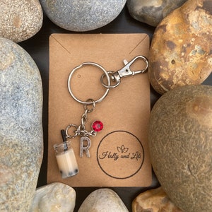 Może przedstawiać: Brązowo-biały brelok do kluczy w kształcie bubble tea z czerwonym kryształowym dodatkiem i przymocowaną literą "R". Brelok znajduje się na brązowej kartce z tekstem "Holly and Lili" w kółku.