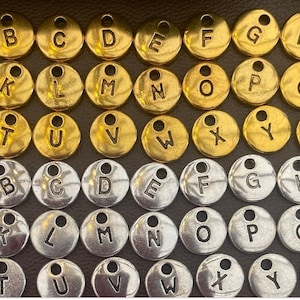 May include: Two sets of alphabet letter charms, one set in gold and one set in silver. Each charm is a small, round disc with a letter printed on it.