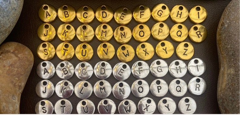 May include: Two sets of alphabet letter charms, one set in gold and one set in silver. Each charm is a small, round disc with a letter printed on it.
