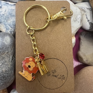 May include: Gold keyring with a lion charm, a red rhinestone, and the letter M. The keyring is attached to a brown card with the text "My and Lita" printed on it.