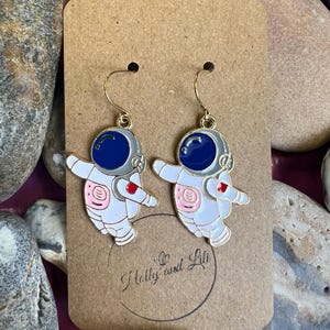 Può includere: Un paio di orecchini a tema astronauta. Ogni orecchino presenta una tuta spaziale bianca con dettagli rosa, un casco blu e ganci dorati. Gli orecchini sono esposti su una scheda marrone con il testo "Holly and Lili".