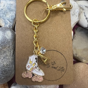 Op de afbeelding: Gouden rolstoelschlüsselhanger met een roze en wit ontwerp, een bliksem en een ster. De sleutelhanger heeft een gouden ketting en een gouden ring met een sluiting. De sleutelhanger bevindt zich op een bruine kaart met de tekst "Folly and Lib" in een cirkel.