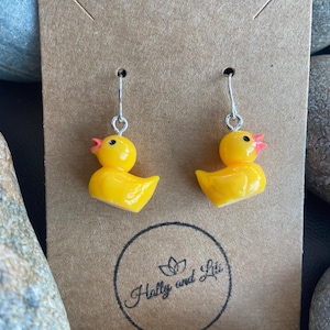 Könnte beinhalten: Ein Paar gelbe Gummiente-Ohrringe mit silbernen Haken. Die Ohrringe befinden sich auf einer braunen Karte mit dem Text "Holly and Liz" in Kursivschrift.