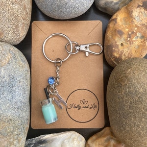 Peut inclure: Un porte-clés argenté avec un charme représentant une petite bouteille en verre contenant un liquide bleu et une paille, un cristal bleu et une lettre H argentée. Le charme est attaché à un anneau argenté et un fermoir à homard argenté. Le porte-clés est sur une carte brune avec le texte "Holly and Lili" dans un cercle.