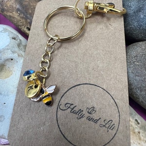 May include: Gold keyring with a bee charm, a blue crystal charm, and a gold initial charm. The keyring is attached to a brown card with the text "Holly and Lili" in a circle.