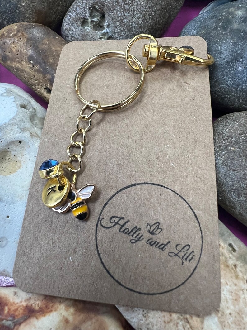 May include: Gold keyring with a bee charm, a blue crystal charm, and a gold initial charm. The keyring is attached to a brown card with the text "Holly and Lili" in a circle.
