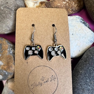 Puede incluir: Un par de pendientes de esmalte negro con forma de mandos de videojuegos. Los pendientes tienen ganchos plateados y detalles de botones de colores. Se exhiben en una tarjeta marrón con el texto "Holly and Lili" en un círculo.
