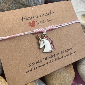 Puede incluir: Una pulsera hecha a mano con un dije de unicornio. El dije es blanco con detalles rosas y un cuerno dorado. La pulsera está ensartada en un cordón rosa y blanco, adjunta a una tarjeta marrón con el texto "Hand made With love" y "DO ALL THINGS WITH LOVE and be powerful, brilliant and brave."