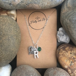 Puede incluir: Un collar de plata con un dije de corazón, un dije de la letra 'R' y una piedra preciosa verde. El collar está sobre una tarjeta marrón con el texto "Holly and Lili" impreso en ella.
