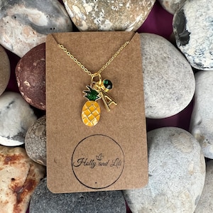 Puede incluir: Un collar de cadena dorada con un dije de piña, una gema verde y un dije de letra 'A' dorada. El collar se muestra en una tarjeta marrón con el texto 'Holly and Lili'.