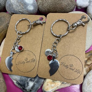 Peut inclure: Deux porte-clés argentés avec un pendentif en forme de demi-cœur et une gemme rouge. Chaque porte-clés est fixé à une carte marron avec le texte "Holly and Lili". Présentés sur une surface rose avec des pierres.