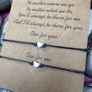 Puede incluir: Dos pulseras de cuerda negra, cada una con un dije de corazón plateado. Las pulseras se exhiben en una tarjeta marrón con el texto "No matter where we go..." y "One for you" y "One for me."