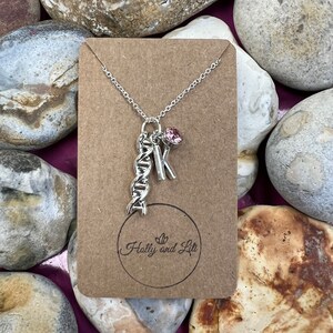 Puede incluir: Collar de plata con un dije de ADN, un dije de la letra K y un cristal rosa. El collar está sobre una tarjeta marrón con el texto "Holly and Lil" en un círculo.