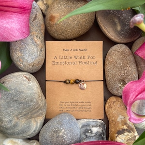 Peut inclure: Une carte brune avec le texte "Make A Wish Bracelet" et "A Little Wish For Emotional Healing". Un bracelet en cordon brun avec une perle en œil de tigre et un charme argenté avec la lettre "R" est sur la carte. La carte est entourée de fleurs roses et de rochers gris.