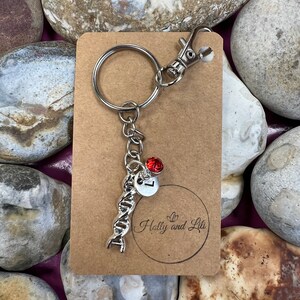 Puede incluir: Un llavero plateado con un dije de hélice de ADN, un strass rojo y un dije plateado con la letra "L". El llavero está unido a una tarjeta marrón con el texto "Holly and Lili" en un círculo.