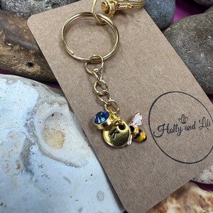 May include: Gold keyring with a bee charm and a blue crystal. The charm has a gold disc with the letter K on it. The keyring is on a brown card with the text "Holly and Lili" on it.