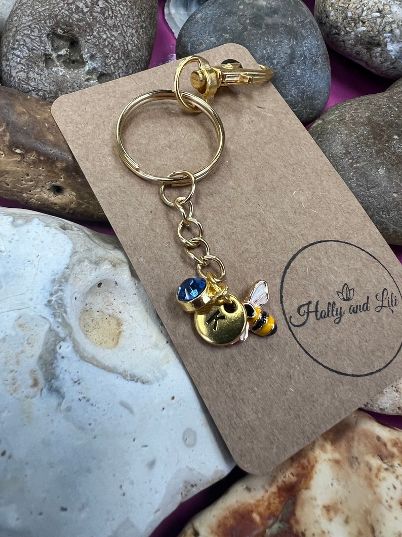May include: Gold keyring with a bee charm and a blue crystal. The charm has a gold disc with the letter K on it. The keyring is on a brown card with the text "Holly and Lili" on it.