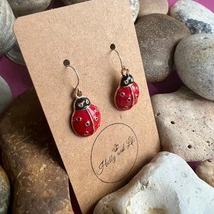May include: A pair of gold ladybug earrings with red and black enamel. The earrings are displayed on a brown card with the text "Holly and Lili" written on it.