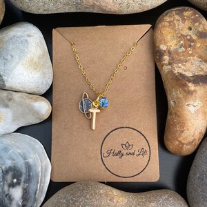 Puede incluir: Collar de oro con un pequeño dije de planeta y un dije de cristal azul. El dije es un planeta dorado con un diseño estrellado en blanco y negro. El collar está sobre una tarjeta marrón con el texto "Holly and Lili" en un círculo.