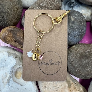 May include: Gold keychain with a small crown charm, a letter E charm, and a pink rhinestone. The keychain is on a brown card with the text "Holly and Liti" in a circle.