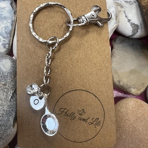 Peut inclure: Un porte-clés en forme de cuillère en argent avec un charme en cristal et la lettre "O" gravée sur la cuillère. Le porte-clés est attaché à un anneau brisé en argent et à une chaîne en argent. Le porte-clés est présenté sur une carte brune avec le texte "Holly and Lita" imprimé dessus.