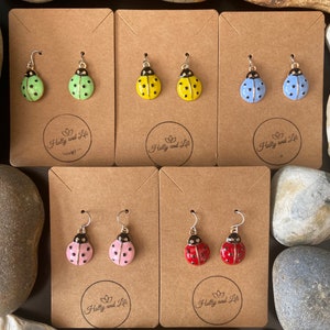 May include: Four pairs of ladybug earrings displayed on cards. The earrings are made of metal and have a gold finish. The ladybugs are painted in different colours: green, yellow, blue, pink, and red. Each card has the text "Holly and Life" printed on it.