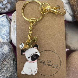 May include: Gold key chain with a white pug charm with black ears and a pink collar. The charm has a rhinestone on the top. The keychain is attached to a brown card with the text "Holly and Lil" in a circle.
