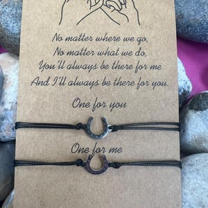 Puede incluir: Dos pulseras negras a juego con dijes de herradura plateados se muestran en una tarjeta marrón. La tarjeta tiene el texto "No matter where we go..." y "One for you, One for me."