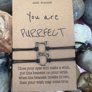 Puede incluir: Dos pulseras de deseos negras con dijes en forma de gato se muestran en una tarjeta marrón. La tarjeta dice "You are PURRFECT" e incluye instrucciones. El fondo presenta varias piedras grises y beige.