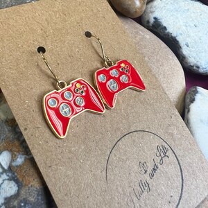 Puede incluir: Pendientes rojos y dorados con forma de mandos de videojuegos. Los pendientes tienen ganchos dorados y se muestran en una tarjeta marrón. La tarjeta tiene el texto "Holly and Lil" en un círculo.