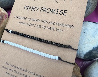 Puede incluir: Dos pulseras de cuentas, una negra y otra blanca, se muestran en una tarjeta con el texto "Pinky Promise". La tarjeta presenta un dibujo lineal de dos manos haciendo una promesa meñique y texto adicional.