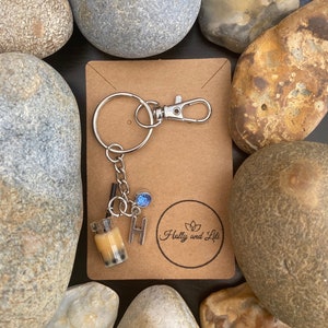 Może przedstawiać: Brązowa kartka z tektury z przymocowanym srebrnym brelokiem. Brelok zawiera małą, przezroczystą plastikową butelkę wypełnioną brązową cieczą i czarnymi perełkami boba. Butelka ma srebrną słomkę i niebieski charm. Na kartce znajduje się również mały srebrny charm z literą "H" i tekstem "Holly and Lili" w kółku.