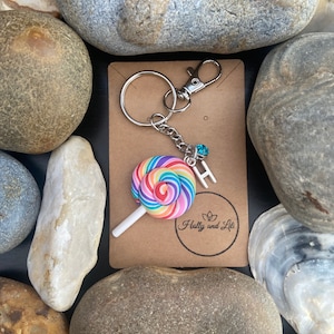 May include: A rainbow swirl lollipop charm attached to a silver key chain with a silver ring and a blue rhinestone. The charm is attached to a brown card with the text "Holly and Lili" printed on it.