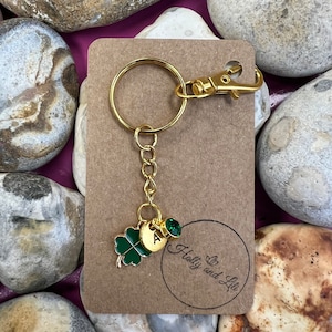 May include: Gold keychain with a green four-leaf clover charm, a gold charm with the letter "A" and a green gemstone. The keychain is on a brown card with the text "Holly and Lily" in a circle.