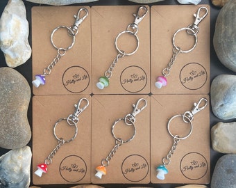 Puede incluir: Seis llaveros plateados con dijes de hongo. Los dijes son de diferentes colores, incluyendo morado, verde, rosa, naranja y azul. Cada llavero tiene una pequeña etiqueta que dice "Holly and Life".