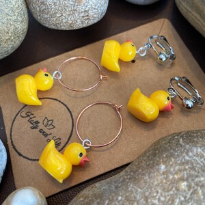 K&ouml;nnte beinhalten: Gelbe Enten-Ohrringe mit Clip-On- und Creolen-Stilen. Die Ohrringe zeigen kleine, gl&auml;nzende gelbe Entenfiguren mit roten Schn&auml;beln und schwarzen Augen. Die Ohrringe werden auf einer braunen Karte mit dem Text "Holly and Lilie" pr&auml;sentiert.