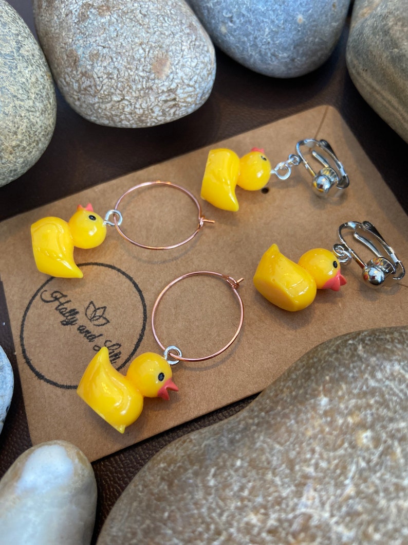 K&ouml;nnte beinhalten: Gelbe Enten-Ohrringe mit Clip-On- und Creolen-Stilen. Die Ohrringe zeigen kleine, gl&auml;nzende gelbe Entenfiguren mit roten Schn&auml;beln und schwarzen Augen. Die Ohrringe werden auf einer braunen Karte mit dem Text "Holly and Lilie" pr&auml;sentiert.