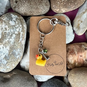 May include: A yellow rubber ducky keyring with a silver charm and a green gemstone. The keyring is attached to a silver key ring. The keyring is on a brown card with the text "Holly and Lili" printed on it.