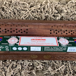 Könnte beinhalten: Ein hölzerner Räucherstäbchenhalter mit einer Schachtel Satya Jasmine Incense. Die Schachtel ist grün und weiß mit dem Schriftzug "Jasmine Incense". Der Halter ist braun mit dekorativen Aussparungen.