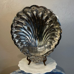 May include: A large silver serving dish shaped like a seashell with a decorative stand. The dish is highly polished and reflects light.