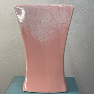 Può includere: Un vaso in ceramica rosa con una glassa bianca. Il vaso ha un'ampia apertura e una base stretta.