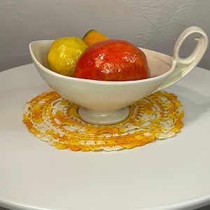 May include: A white ceramic serving bowl with a handle, filled with a red and yellow fruit, sitting on a yellow and white crocheted doily.