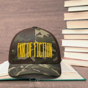 May include: A camouflage trucker hat with the text "FAN OF FICTION" embroidered in yellow. The hat is resting on an open book, with a stack of books in the background. The hat has a brown mesh back.
