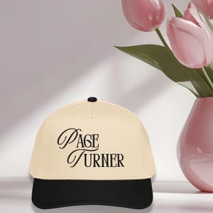 May include: A beige baseball cap with a black brim and a black button on top. The words "Page Turner" are written in elegant black script across the front of the cap. The cap is on a white surface.
